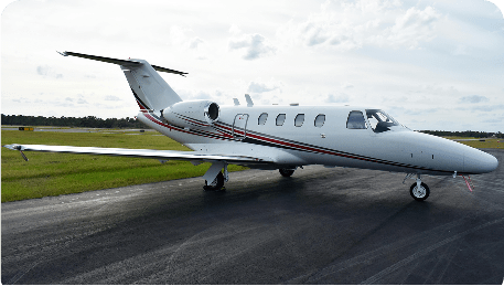 A white Cessna Citation light business jet parked on a dark asphalt runway. The aircraft features a clean red and grey stripe along the fuselage under a bright, overcast sky with a line of trees in the background.