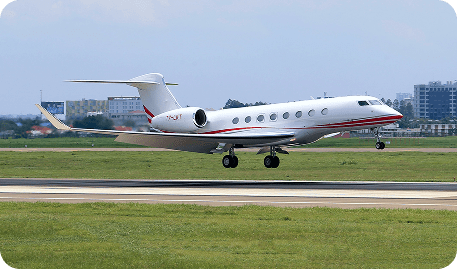 A white private jet with black racing stripes parked on a wet runway at twilight. The sky is a mix of deep purple and orange clouds, with the plane’s navigation lights casting a warm glow on the asphalt.