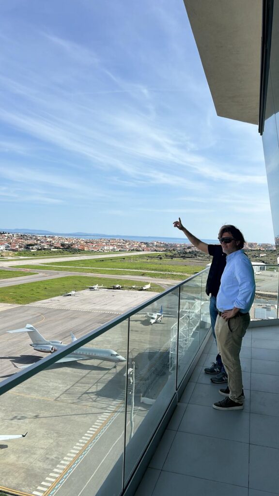 Wide shot from a modern balcony with glass railings. A man in a light blue shirt and khakis looks on as another man points toward an airport runway. On the tarmac below, a large silver private jet is parked. In the far distance, a city coastline meets the sea under a streaky blue sky.