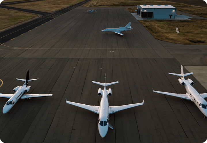 High-angle aerial view of three white private jets parked side-by-side on an asphalt tarmac. A fourth jet is parked further back near a small hangar, all captured under the soft, warm light of late afternoon.
