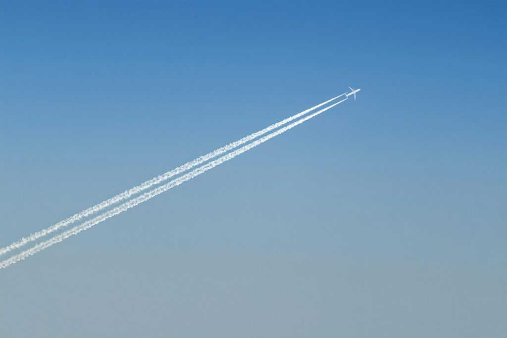 A small silhouette of a jet soaring through an expansive, cloudless blue sky, trailing two parallel white lines that stretch across the horizon.